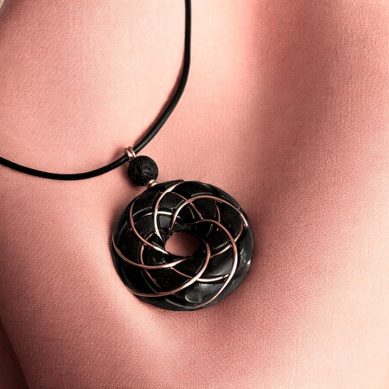 May include: A black pendant necklace with a circular design featuring a rose gold wire pattern. The pendant is hanging on a black cord.