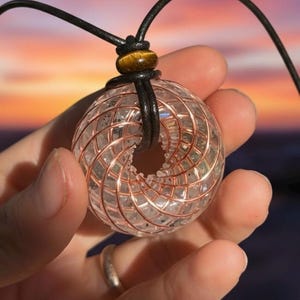 May include: A clear quartz crystal pendant with copper wire detailing, suspended from a black leather cord. The pendant has a spherical shape with a central hole and a tiger's eye bead accent. The background shows a colorful sunset.