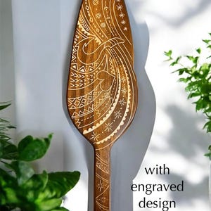 Op de afbeelding: Een houten peddel met een ingewikkeld gegraveerd ontwerp. Het ontwerp heeft een golfpatroon met geometrische vormen en een gestileerde vogel.