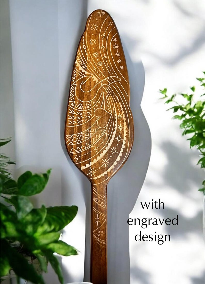 Op de afbeelding: Een houten peddel met een ingewikkeld gegraveerd ontwerp. Het ontwerp heeft een golfpatroon met geometrische vormen en een gestileerde vogel.