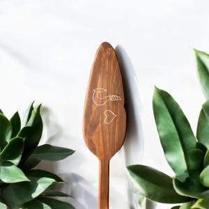Op de afbeelding: Een houten spatel met een bruine vlek en een wit ontwerp van een haak en een hart. De spatel heeft een lange steel.