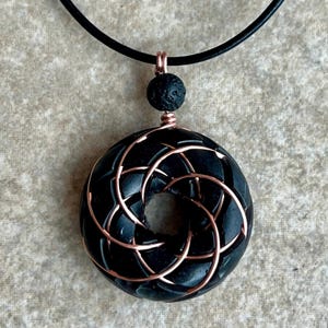 May include: A black lava rock pendant with a copper wire design, hanging from a black leather cord.
