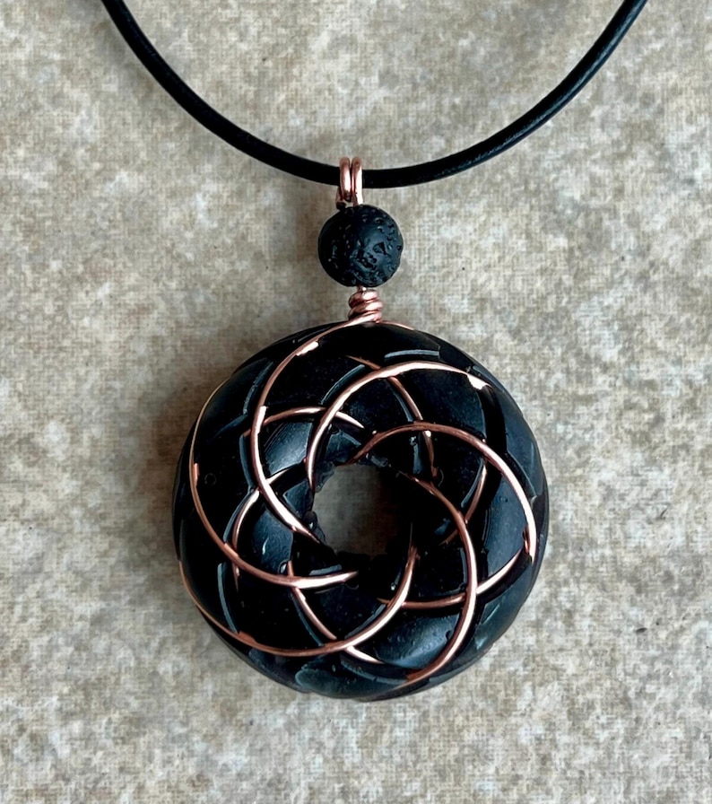 May include: A black lava rock pendant with a copper wire design, hanging from a black leather cord.