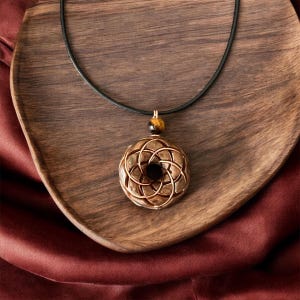 May include: A brown wooden pendant with a geometric design hangs from a black leather cord. The pendant is accented with a tiger's eye stone.