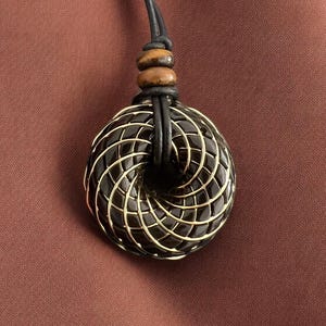 May include: A black and gold geometric pendant necklace with a leather cord. The pendant is a circular design with a spiral pattern.