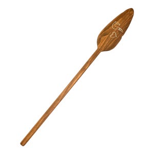 Op de afbeelding: Een houten peddel met een gesneden ontwerp van een vogel en twee harten. De peddel is bruin en heeft een lange steel.