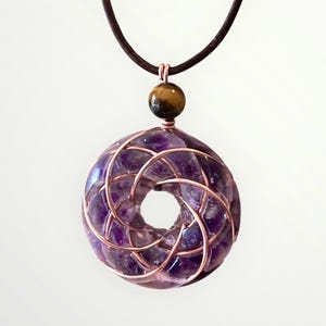 May include: A handcrafted pendant necklace featuring a donut-shaped amethyst stone encased in copper wire. A small tiger's eye bead hangs above the amethyst, suspended from a brown leather cord. The amethyst is a deep purple color.