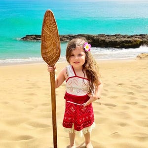 Op de afbeelding: Een jong meisje verkleed als Moana, draagt een rode en witte jurk en houdt een houten peddel vast met ingewikkelde houtsnijwerk. Ze staat op een zandstrand met de oceaan op de achtergrond.