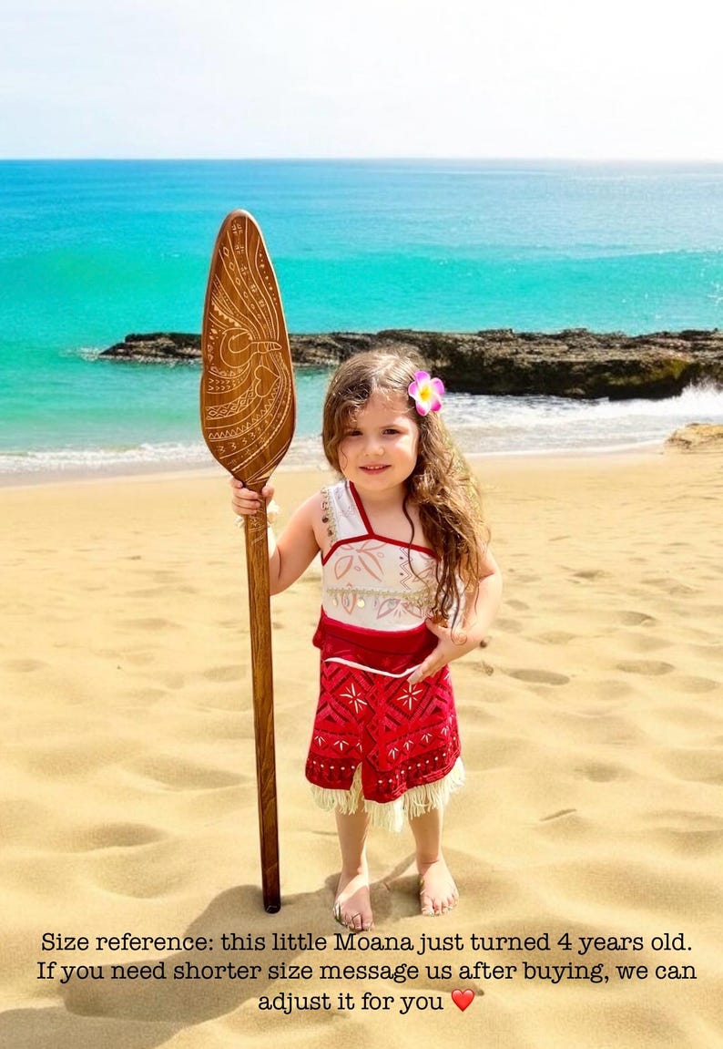 Op de afbeelding: Een jong meisje verkleed als Moana, draagt een rode en witte jurk en houdt een houten peddel vast met ingewikkelde houtsnijwerk. Ze staat op een zandstrand met de oceaan op de achtergrond.