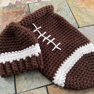 May include: A brown and white crocheted football hat and a matching football-shaped blanket. The hat has a white stripe around the bottom and the blanket has a white stripe around the edge.