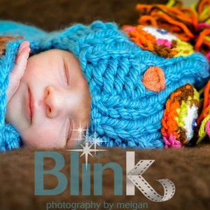 Puede incluir: Un bebé durmiendo con un gorro azul tejido con detalles de ganchillo de colores. El gorro tiene un gran pompón de hilo naranja, amarillo y rosa.
