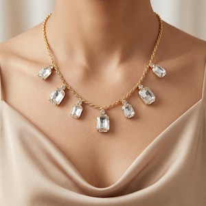 May include: A gold-coloured necklace with a rope chain design, adorned with multiple clear, rectangular crystal pendants. The crystals are evenly spaced along the chain, creating a sparkling, elegant design. A statement necklace.