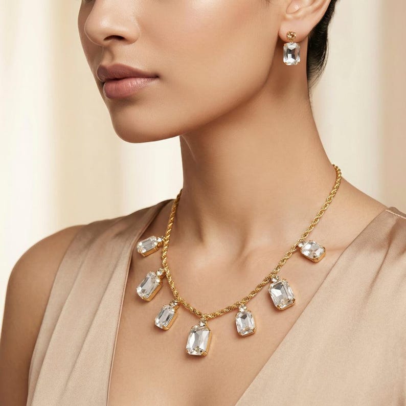 May include: A gold-toned necklace and matching earrings set. The necklace features a rope chain with multiple rectangular, clear crystal pendants. The earrings have a single rectangular crystal. The jewelry is worn by a person wearing a beige top.