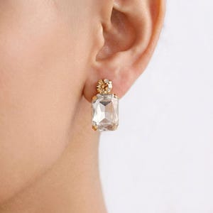 May include: A close-up of a gold-toned earring with a large, rectangular, clear crystal. The earring has a smaller, round crystal at the top. The earring is on a person's ear.
