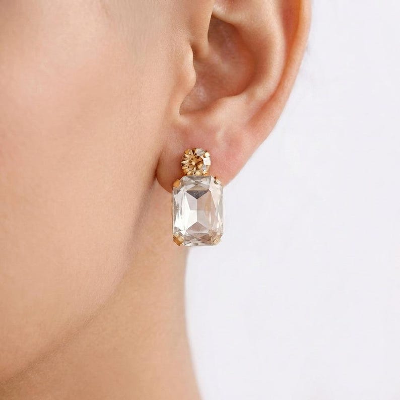 May include: A close-up of a gold-toned earring with a large, rectangular, clear crystal. The earring has a smaller, round crystal at the top. The earring is on a person's ear.