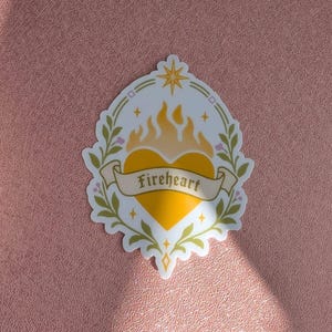 May include: A sticker featuring a yellow heart with flames on top, surrounded by a green leaf wreath and a star at the top. A banner across the heart displays the text Fireheart. The sticker is placed on a textured, metallic rose-gold notebook cover.