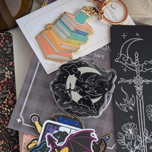 Moon and Dragons Acrylic Kindle Grip. A collection of stationery items including a gold-toned metal keychain featuring a stack of colourful books, a black rectangular bookmark with a white line art sword and floral design.