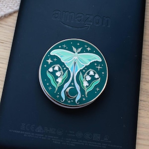 May include: A circular phone grip featuring a dark teal background with a central light green luna moth illustration, white lily of the valley flowers, and a small silver crescent moon. The design includes scattered white stars and sparkles.