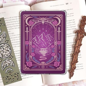 Może przedstawiać: Prostokątne fioletowe etui na czytnik e-booków z górskim krajobrazem, różami i napisem To the stars who listen, w towarzystwie zielonej zakładki z fazami księżyca oraz drewnianej zakładki w kształcie miecza, leżące na otwartej książce.