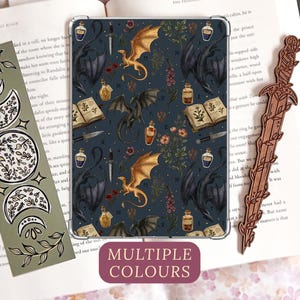 Puede incluir: Una funda rectangular para tableta con fondo azul oscuro y un patrón de dragones dorados y negros, libros abiertos, dagas y frascos de pociones. A la izquierda, un marcapáginas verde con fases lunares y a la derecha, uno de madera con forma de espada.