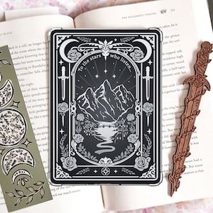 May include: A rectangular black and white e-reader case featuring a mountain landscape, a river, and the text To the stars who listen, flanked by a sage green bookmark with moon phases and a brown wooden sword-shaped bookmark on an open book.