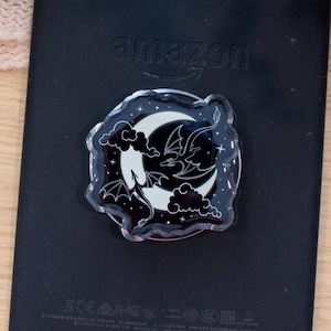 May include: A black phone grip featuring a circular acrylic topper with a white crescent moon, clouds, and a dragon silhouette against a black background with small white stars. The grip is attached to the back of a black e-reader with the word amazon.