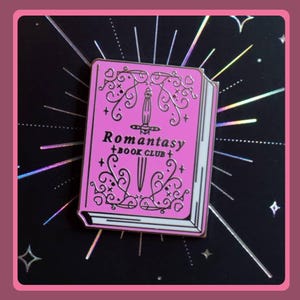 Può includere: Una spilla smaltata rettangolare a forma di libro con copertina rosa brillante, caratterizzata da un'illustrazione centrale di un pugnale argentato, volute decorative e il testo Romantasy Book Club stampato in carattere scuro sulla copertina.