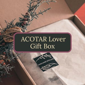 Puede incluir: Una caja de regalo de cartón que contiene papel de seda blanco y una rama de vegetación seca con pequeñas bayas rojas. Se ve una pegatina negra con el texto HAND MADE WITH LOVE y una ilustración de una mano. La caja muestra el texto ACOTAR Lover Gift Box.