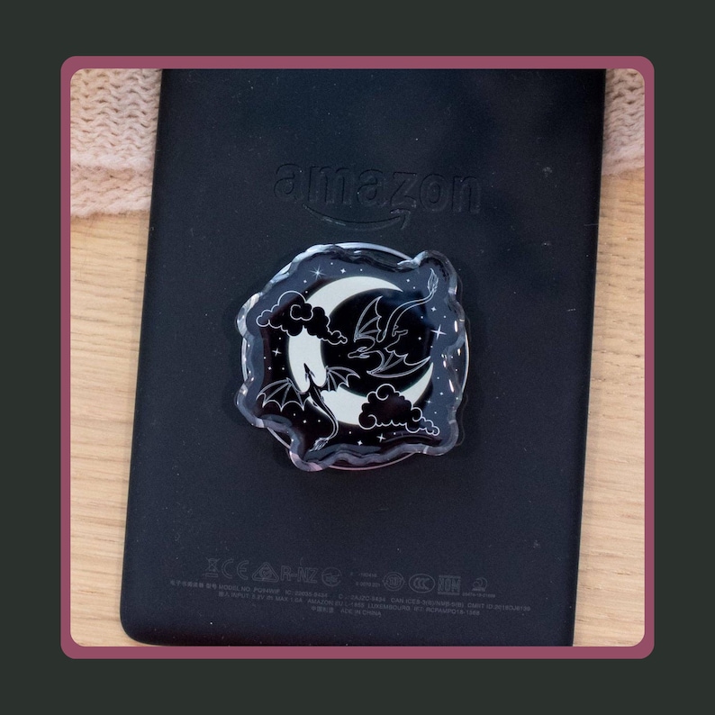 Moon and Dragons Acrylic Kindle Grip. A black phone grip featuring a circular acrylic topper with a white crescent moon, clouds, and a dragon silhouette against a black background with small white stars.