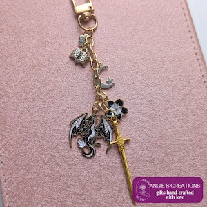 May include: A gold-toned metal keyring featuring multiple charms including a silver open book, a silver crescent moon with a star, a black and white dragon, a black and white flower, and a gold sword. The keyring has a lobster clasp attachment.