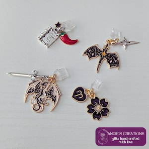 Puede incluir: Cuatro colgantes de pendientes con temática de fantasía. Incluye un libro con el texto "Once Upon A Time", un chile rojo, un dragón, una estrella, una espada, un corazón con el número 3 y una flor negra. Cada colgante tiene una anilla dorada.