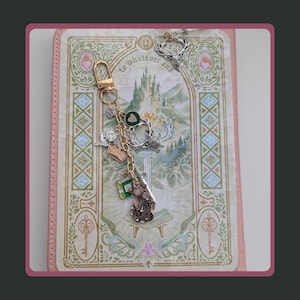 Stag Throne of Glass Inspired Charm Keychain. A gold-toned metal keychain featuring multiple charms including a green heart, a silver crown, a small green book, a sword, and a detailed stag head.