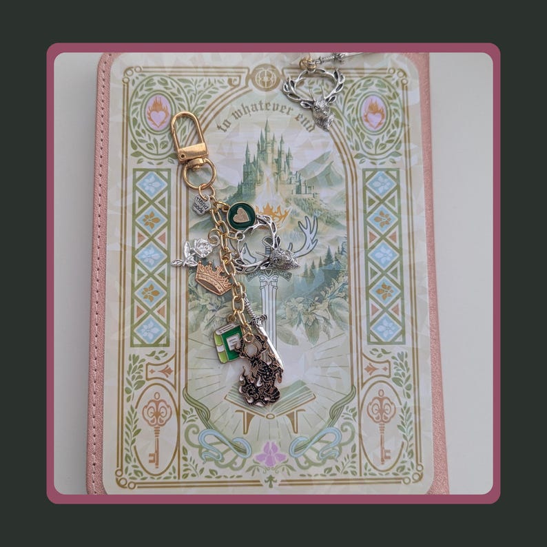 Stag Throne of Glass Inspired Charm Keychain. A gold-toned metal keychain featuring multiple charms including a green heart, a silver crown, a small green book, a sword, and a detailed stag head.