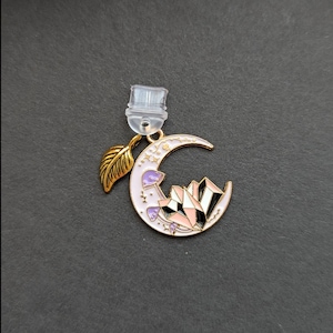 May include: A gold-toned earring featuring a crescent moon design with a light pink enamel finish. The moon is adorned with a cluster of crystals, mushrooms, and stars. A gold leaf charm and a clear earring back complete the design.