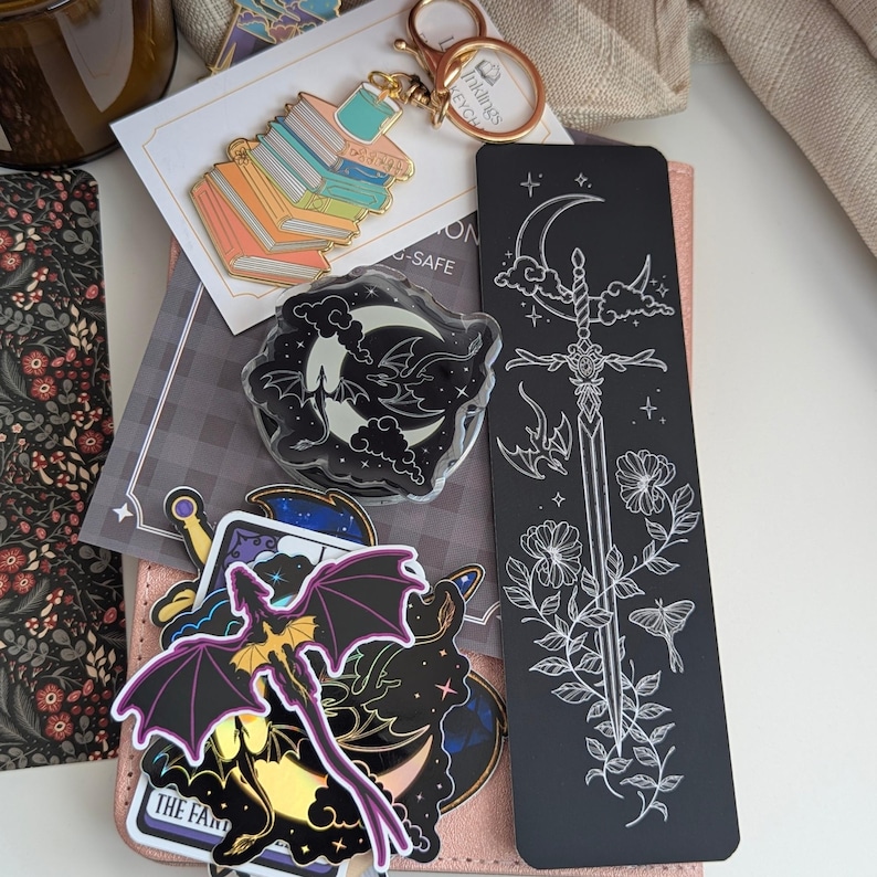 Moon and Dragons Acrylic Kindle Grip. A collection of stationery items including a gold-toned metal keychain featuring a stack of colourful books, a black rectangular bookmark with a white line art sword and floral design.