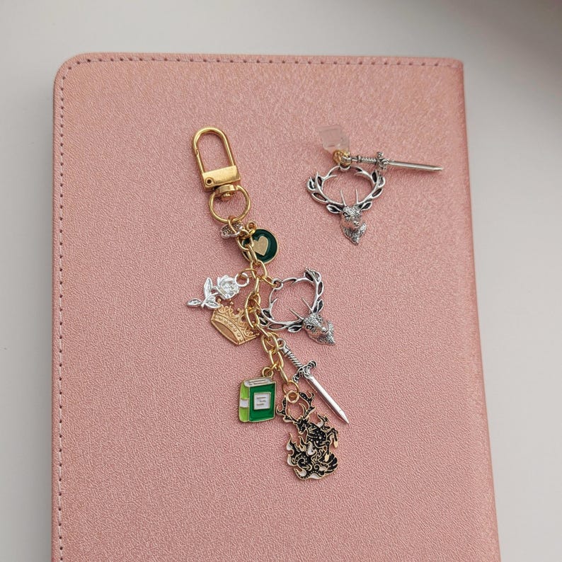 Stag Throne of Glass Inspired Charm Keychain. A gold-toned keyring featuring multiple charms including a green book, a crown, a rose, a heart, a sword, and two silver-toned deer heads.