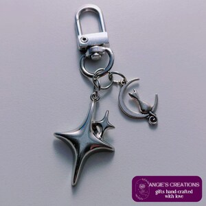 May include: Silver-coloured keychain featuring a starburst charm and a crescent moon charm with a cat silhouette. The keychain includes a lobster clasp and a ring for attaching keys. The text "ANGIE'S CREATIONS gifts hand-crafted with love" is visible.