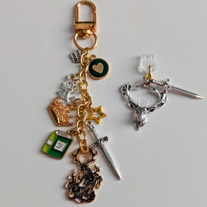 Stag Throne of Glass Inspired Charm Keychain. A gold-toned keyring featuring multiple charms including a green book, a gold crown, a silver rose, a gold star, a silver sword, and a black stag silhouette.