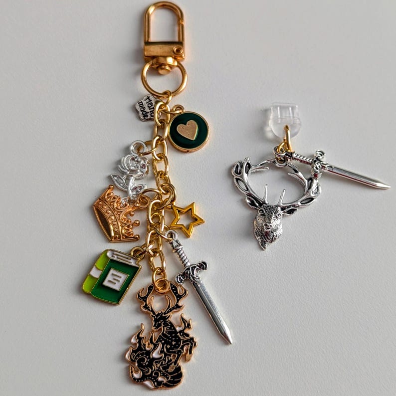Stag Throne of Glass Inspired Charm Keychain. A gold-toned keyring featuring multiple charms including a green book, a gold crown, a silver rose, a gold star, a silver sword, and a black stag silhouette.