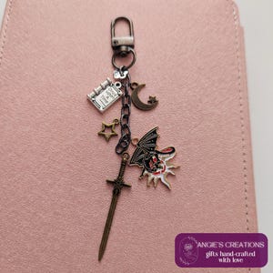 May include: A keychain with various charms, including a book with "Once upon a Time" text, a crescent moon, a star, a sword, and a sun with a bat-like creature. The charms are attached to a silver-colored clasp.