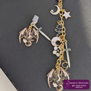 May include: Gold-toned earrings featuring dragon and sword charms, along with moon, star, and floral accents. The earrings have a dangling design with intricate details and a fantasy theme. The text "ANGIE'S CREATIONS gifts hand-crafted with love" is visible.