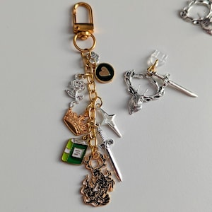 Stag Throne of Glass Inspired Charm Keychain. A gold-toned metal keyring featuring a lobster clasp and a gold chain adorned with various charms including a silver rose, a gold crown, a green book, a silver sword, a four-pointed star.