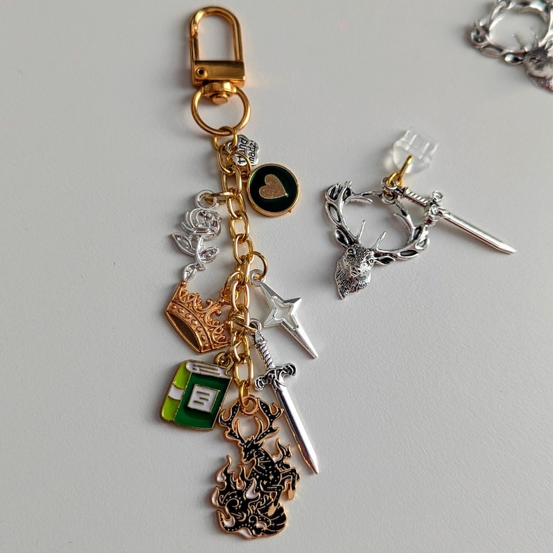 Stag Throne of Glass Inspired Charm Keychain. A gold-toned metal keyring featuring a lobster clasp and a gold chain adorned with various charms including a silver rose, a gold crown, a green book, a silver sword, a four-pointed star.