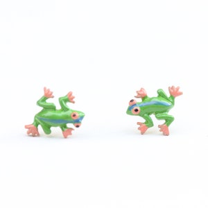 May include: Two small, green frog figurines with pink feet and red eyes are displayed against a white background. Each frog has a blue stripe down its back. The frogs are positioned facing each other.