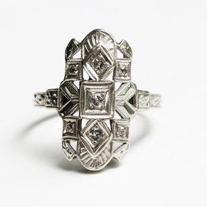 May include: A silver ring with a geometric design featuring multiple small diamonds. The ring has a delicate, intricate design.