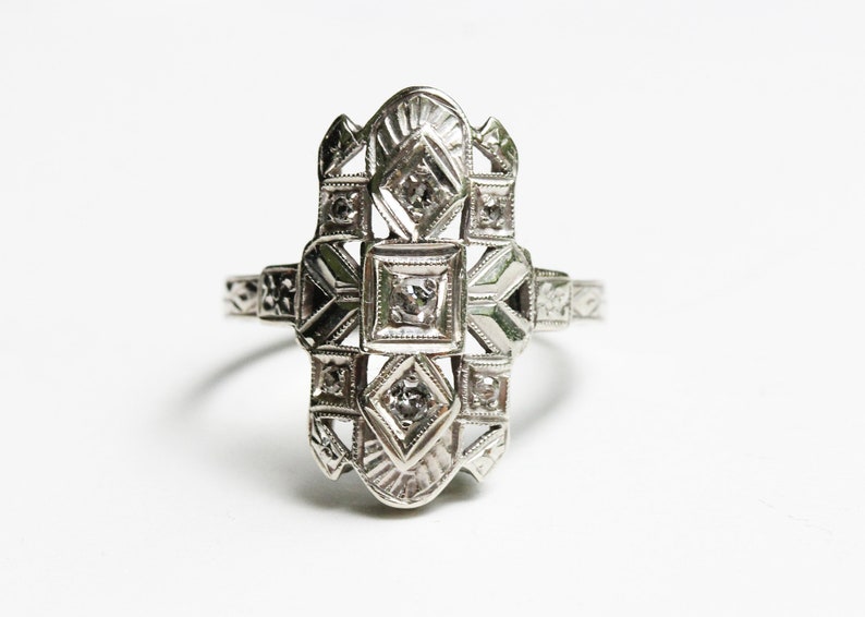 May include: A silver ring with a geometric design featuring multiple small diamonds. The ring has a delicate, intricate design.