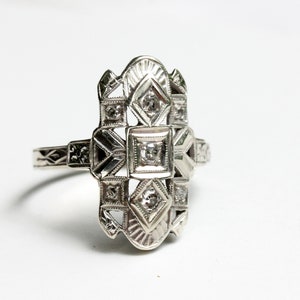 May include: A silver ring with a geometric design featuring multiple small diamonds. The ring has a delicate, intricate design.