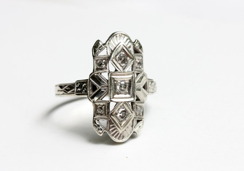 May include: A silver ring with a geometric design featuring multiple small diamonds. The ring has a delicate, intricate design.