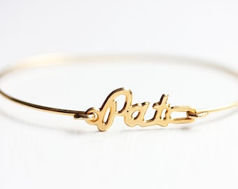 May include: A gold-colored bangle bracelet with the name "Pat" in a cursive font. The bracelet is thin and circular, with a clasp closure. The name is attached to the bracelet.
