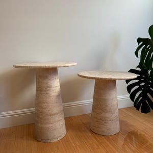 May include: Two beige stone side tables with round tops and conical bases. The tables are made of a light-coloured stone with natural veining. The tables are of different sizes and are placed on a wooden floor.